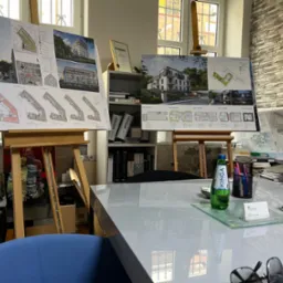 Prezentacja projektów architektonicznych na sztalugach w biurze, widoczne rzuty mieszkań i wizualizacje budynków wielorodzinnych, na stole butelka wody, wizytówka i okulary.