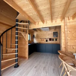 Wnętrze drewnianego domu z spiralnymi schodami, czarną zabudową kuchenną i wiklinowymi krzesłami, oświetlone ciepłym światłem.