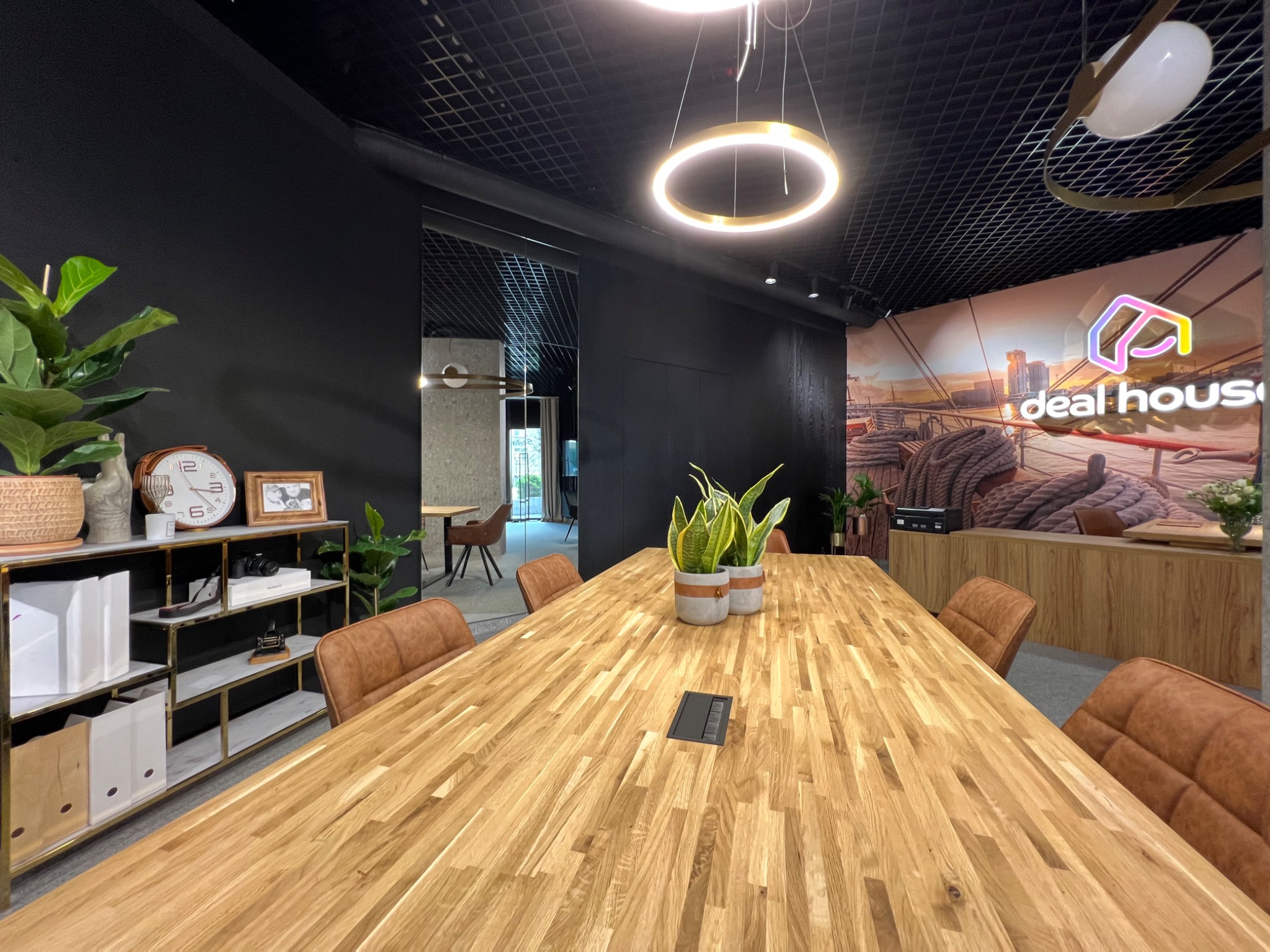 Nowoczesna sala konferencyjna z dużym drewnianym stołem, skórzanymi krzesłami, czarnymi ścianami i designerskim oświetleniem, z logo 'deal house' na ścianie w tle.