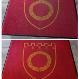 Czerwona wycieraczka wejściowa z żółtym herbem i napisem 'VESTE' na tle szarej podłogi.