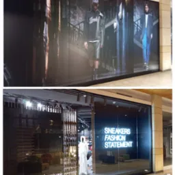 Witryna sklepowa z neonowym napisem 'SNEAKERS FASHION STATEMENT', za szybą widoczne schody, manekin w białym ubraniu i dekoracje.