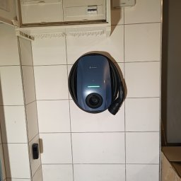 ELEKTRYCY TRÓJMIASTO SPÓŁKA Z OGRANICZONĄ ODPOWIEDZIALNOŚCIĄ - Niebieska stacja ładowania pojazdów elektrycznych Solplanet zamontowana na kafelkowanej ścianie, obok skrzynki bezpiecznikowej i licznika energii.