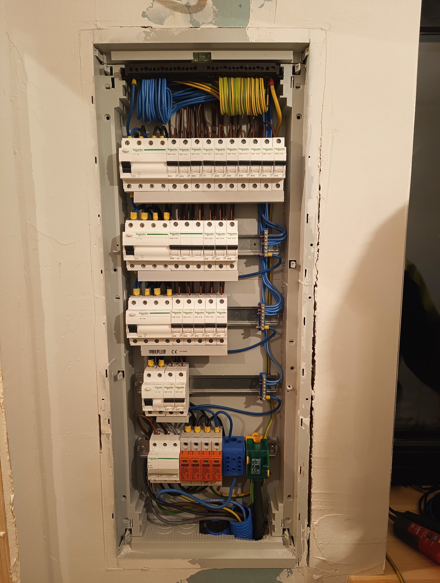 Rozdzielnica elektryczna Schneider w szarej obudowie, z widocznymi bezpiecznikami, przewodami w kolorach niebieskim, brązowym i żółto-zielonym, zamontowana w ścianie.