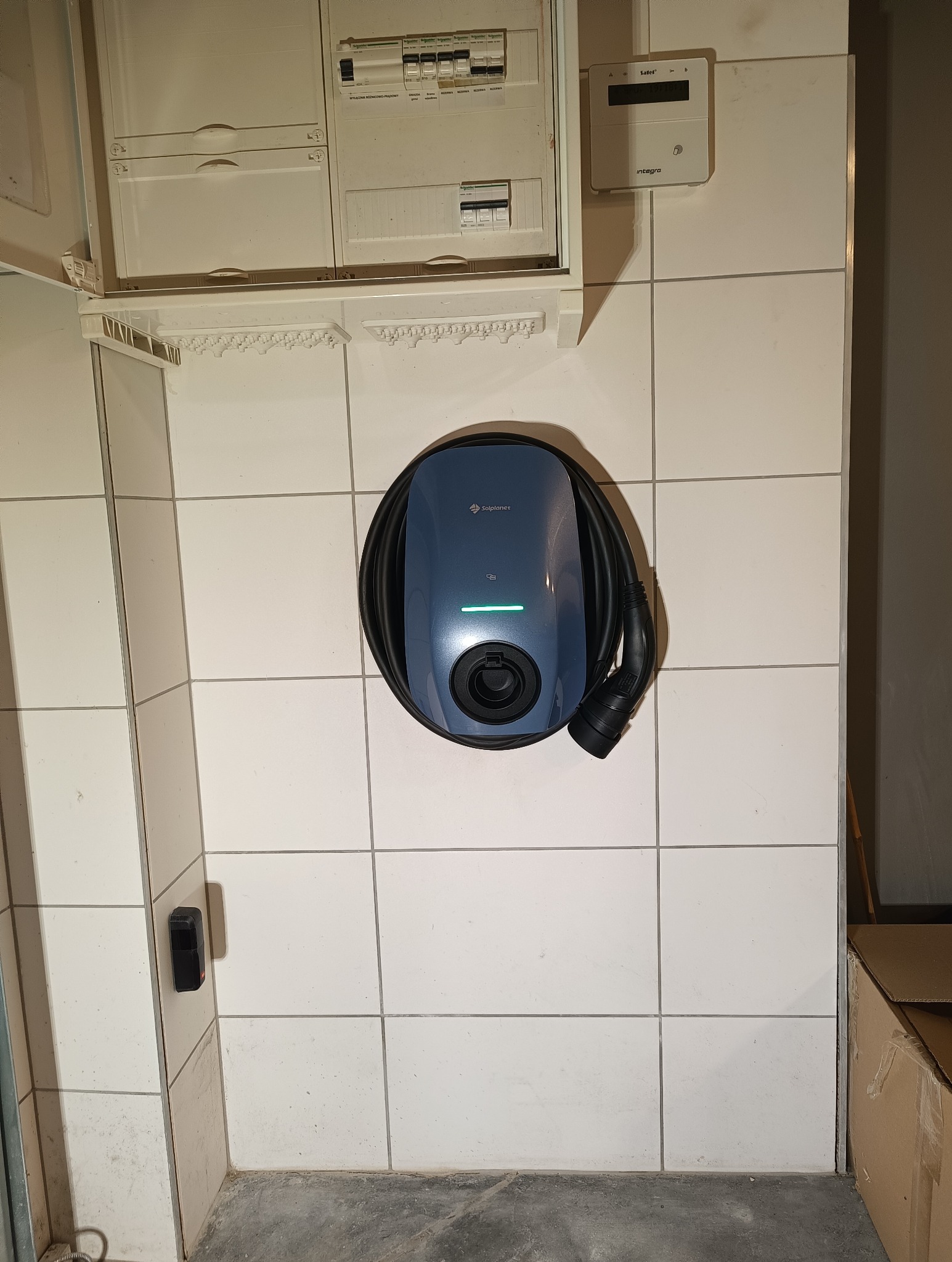 Niebieska stacja ładowania pojazdów elektrycznych Solplanet zamontowana na kafelkowanej ścianie, obok skrzynki bezpiecznikowej i licznika energii.