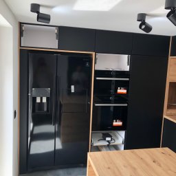 Meble na wymiar Damian Ratajczak - Nowoczesna kuchnia z czarnymi frontami szafek, zabudowaną lodówką side-by-side i piekarnikami, oświetleniem punktowym oraz blatem z jasnego drewna.