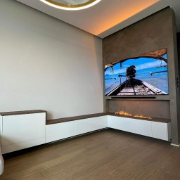 Meble na wymiar Damian Ratajczak - Nowoczesny salon z biało-drewnianą zabudową meblową, biokominkiem pod telewizorem i okrągłą lampą sufitową.