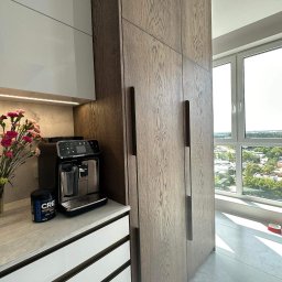 Meble na wymiar Damian Ratajczak - Nowoczesna kuchnia z drewnianymi szafkami sięgającymi sufitu, zintegrowanymi z zabudową, ekspres do kawy i bukiet kwiatów na blacie, widok z okna na miasto.