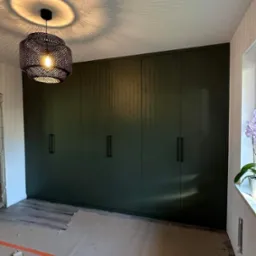 Wnętrze pokoju z dużą, ciemnozieloną szafą wnękową z minimalistycznymi uchwytami, widoczne zabezpieczenia podłogi podczas montażu, oraz ażurowa lampa sufitowa rzucająca wzory na sufit.