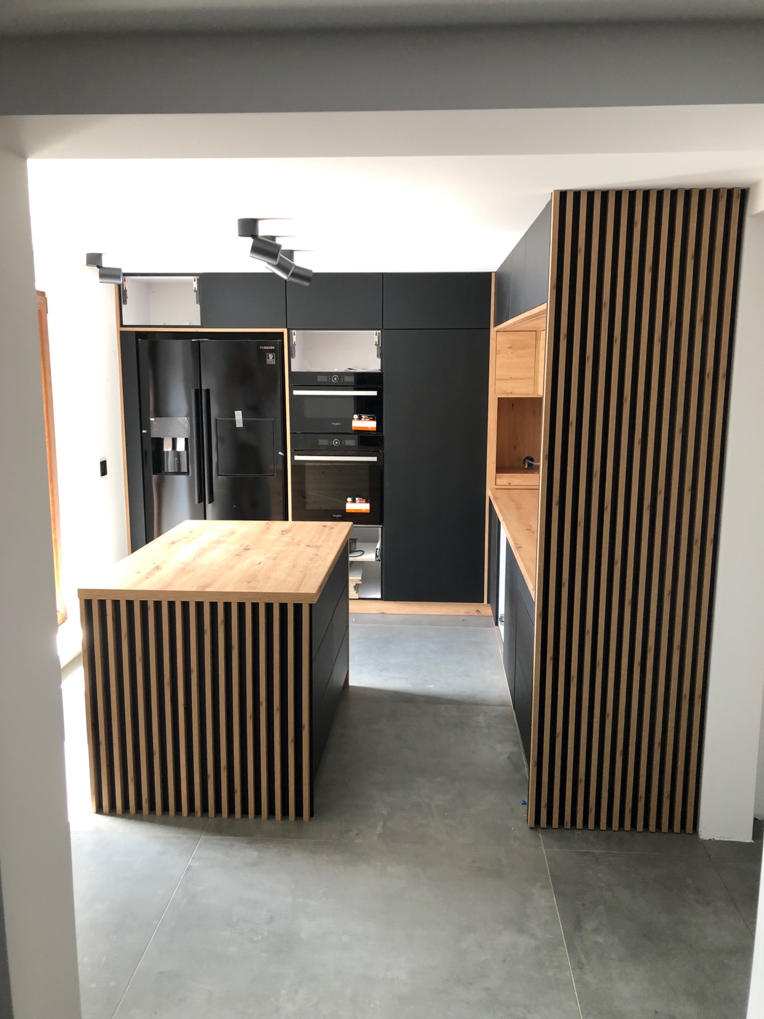 Nowoczesna kuchnia z czarnymi szafkami i wyspą z drewnianymi lamelami; otwarte szafki górne, wbudowane piekarniki i lodówka side-by-side.