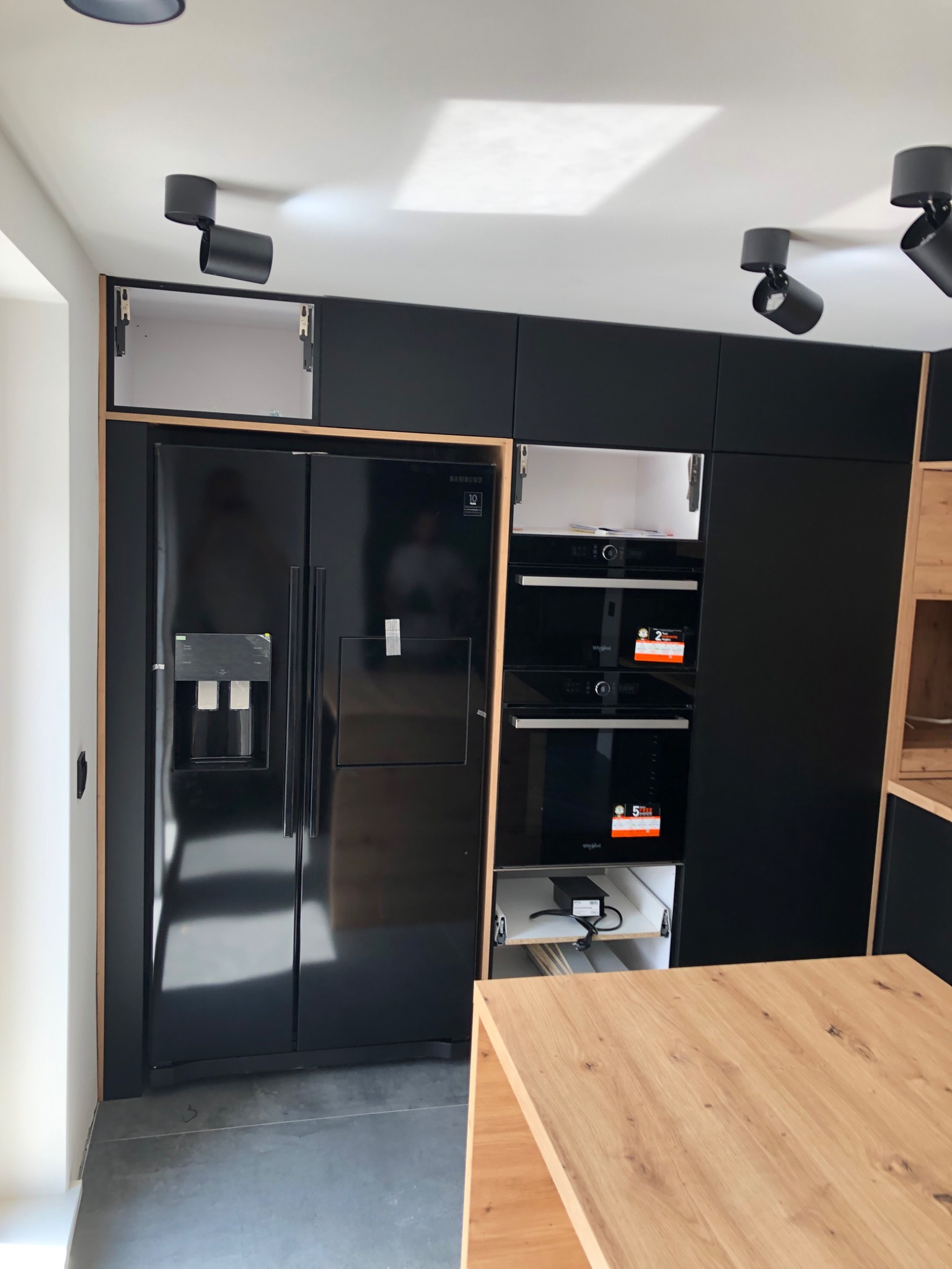 Nowoczesna kuchnia z czarnymi frontami szafek, zabudowaną lodówką side-by-side i piekarnikami, oświetleniem punktowym oraz blatem z jasnego drewna.