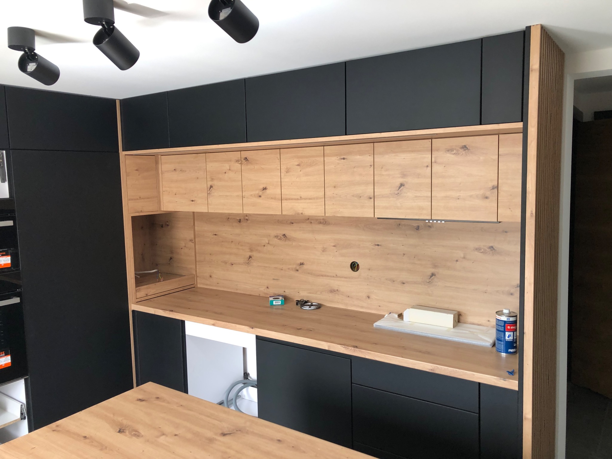 Nowoczesna kuchnia na wymiar z czarnymi szafkami górnymi i dolnymi oraz drewnianym blatem i szafkami wiszącymi, widoczne akcesoria montażowe na blacie.