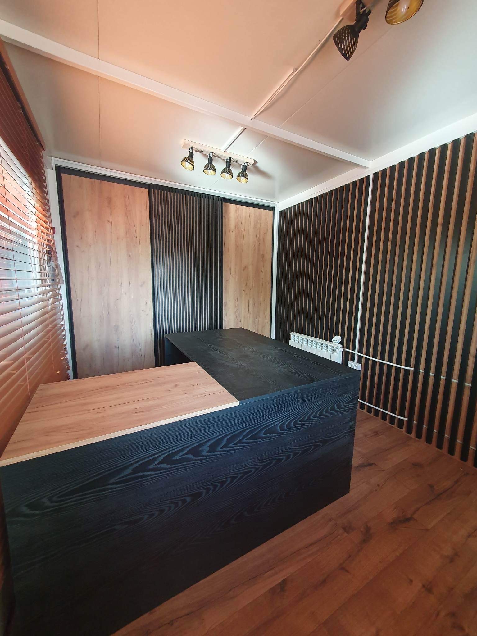 Nowoczesne biuro z czarnym, drewnianym biurkiem na wymiar, uzupełnionym o jasny blat, na tle ściany z drewnianych listew i żaluzji w oknie.