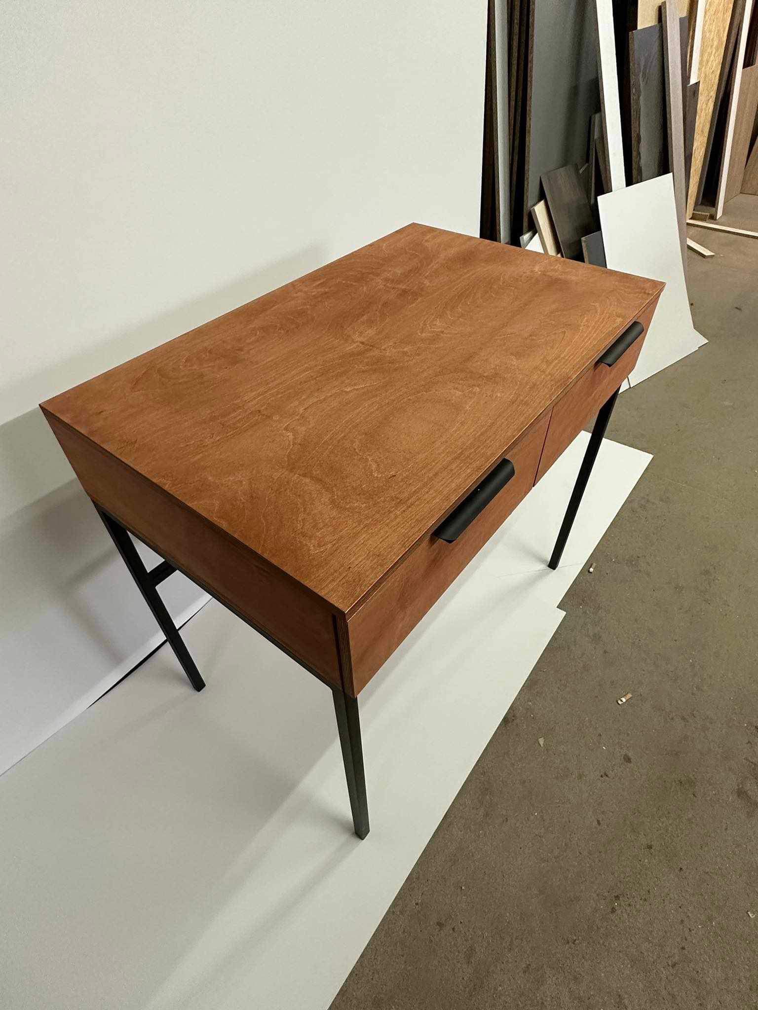 Biurko z drewna o ciepłym odcieniu z czarnymi, metalowymi nogami w kształcie litery X i minimalistycznymi uchwytami szuflad, sfotografowane w warsztacie.