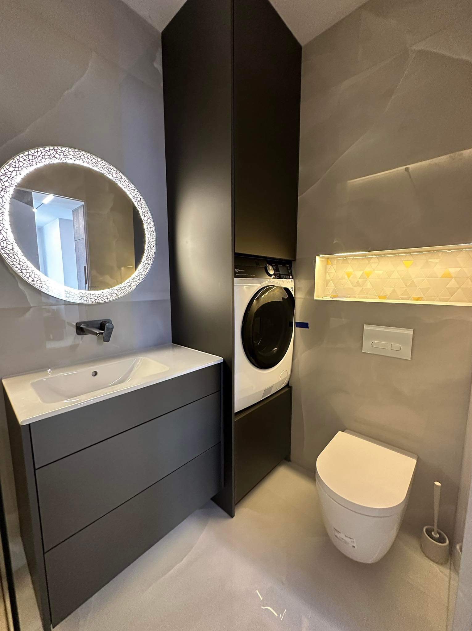 Nowoczesna łazienka w szarościach z okrągłym lustrem LED, zabudowaną pralką i wiszącą toaletą.