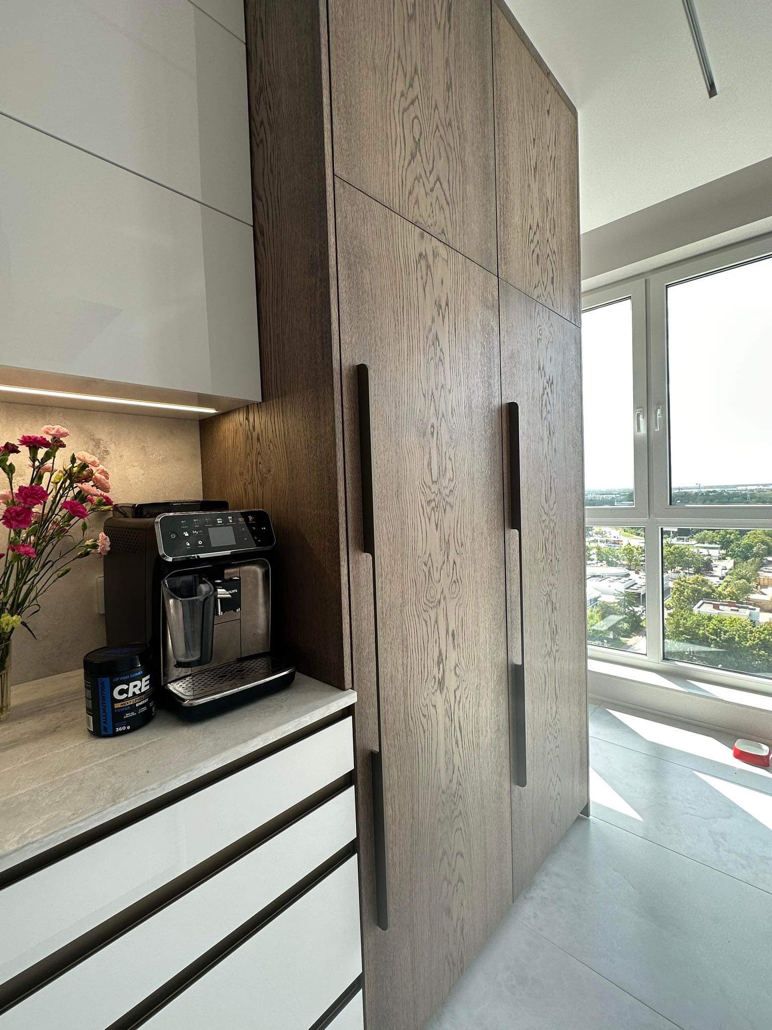 Nowoczesna kuchnia z drewnianymi szafkami sięgającymi sufitu, zintegrowanymi z zabudową, ekspres do kawy i bukiet kwiatów na blacie, widok z okna na miasto.