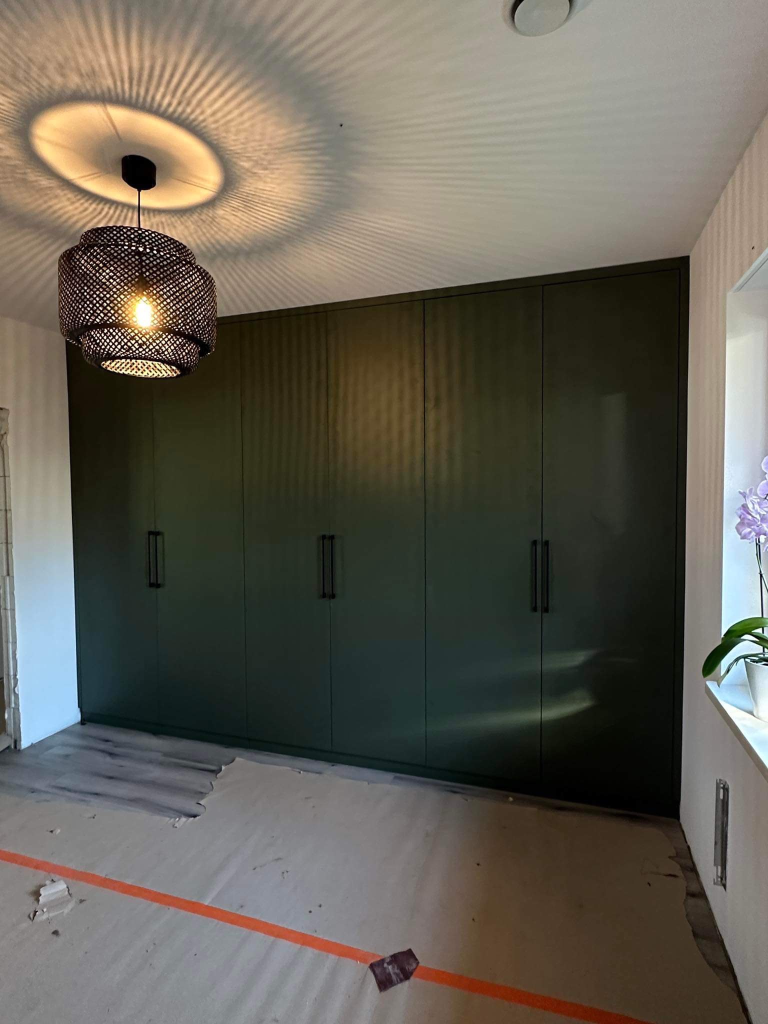 Wnętrze pokoju z dużą, ciemnozieloną szafą wnękową z minimalistycznymi uchwytami, widoczne zabezpieczenia podłogi podczas montażu, oraz ażurowa lampa sufitowa rzucająca wzory na sufit.