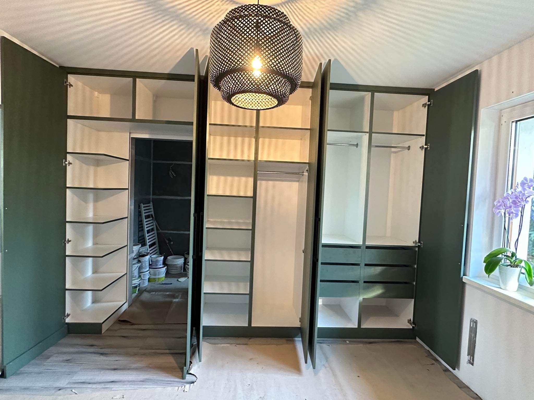 Wnętrze pokoju z dużą, otwartą szafą wnękową z zielonymi drzwiami i białymi półkami, widoczne puszki z farbą i drabina w tle, nad szafą wisi ażurowa lampa.