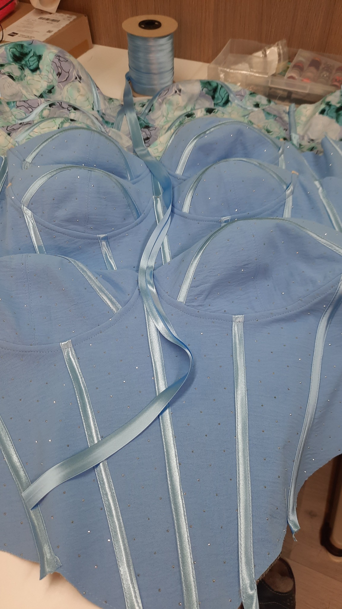 Niebieskie, błyszczące gorsety w trakcie szycia, z satynowymi tasiemkami i rolką tasiemki w tle.