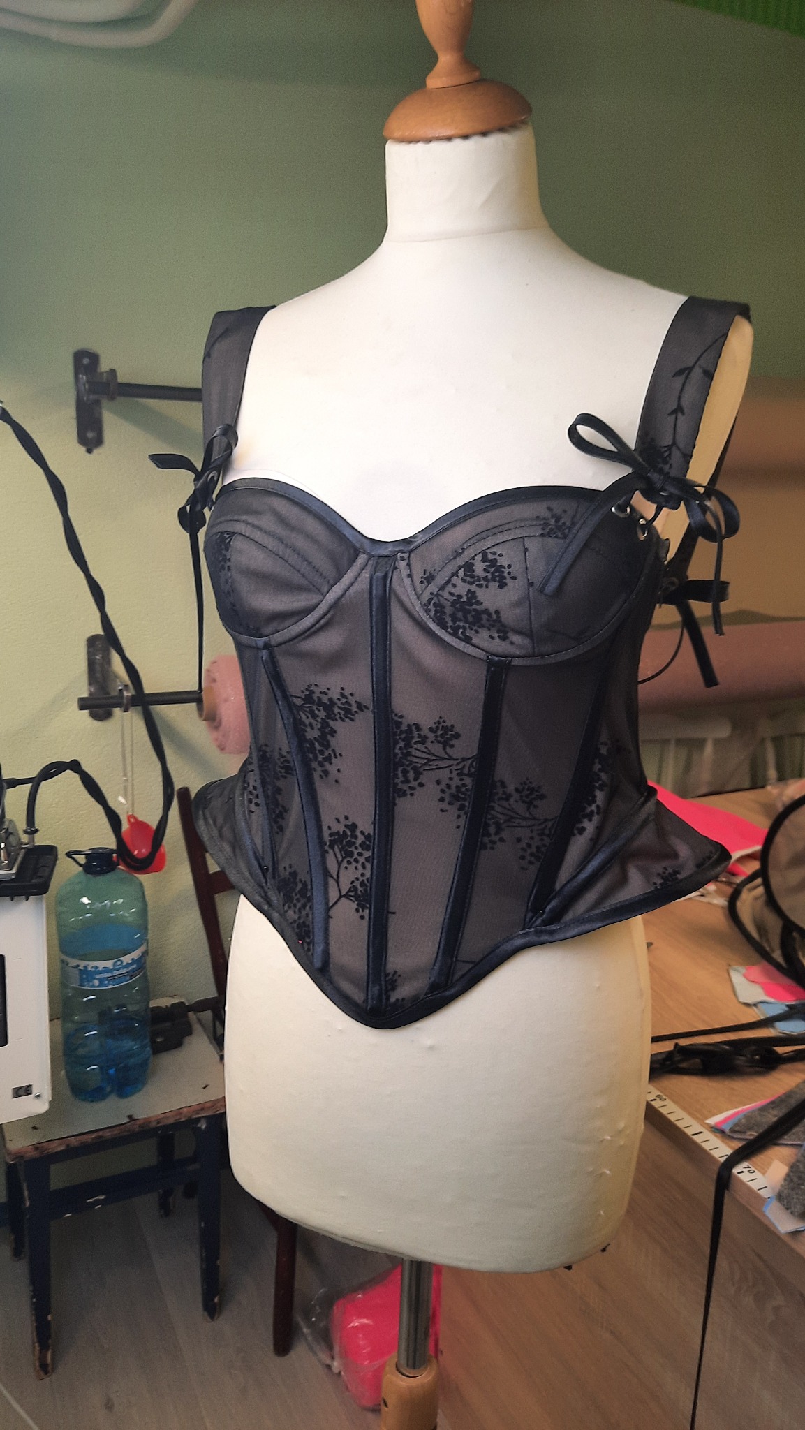 Czarny, koronkowy gorset z motywem gałązek na manekinie krawieckim, widoczne detale wykończenia i wiązania na ramiączkach, w tle warsztat krawiecki.