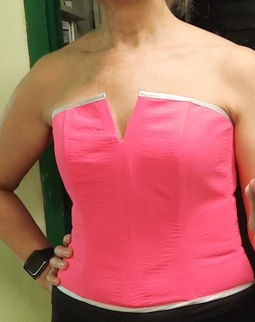 Kobieta pozuje w różowym gorsecie bez ramiączek z srebrnymi detalami, widoczny zegarek na nadgarstku i fragment zielonych drzwi w tle.