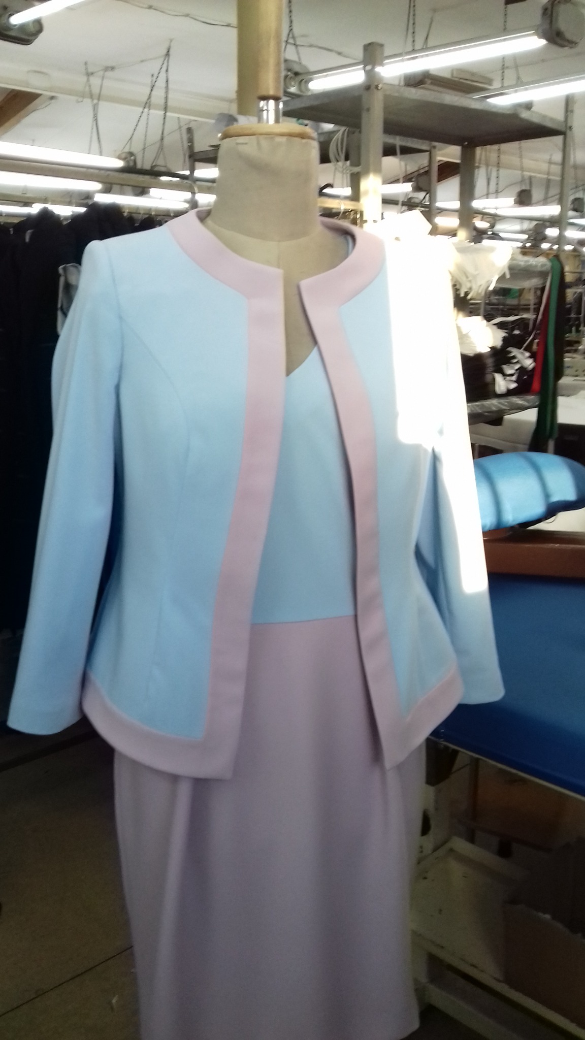 Jasnoniebieski żakiet z różowymi lamówkami i dopasowana sukienka na manekinie w szwalni, widoczne wyposażenie warsztatu w tle.