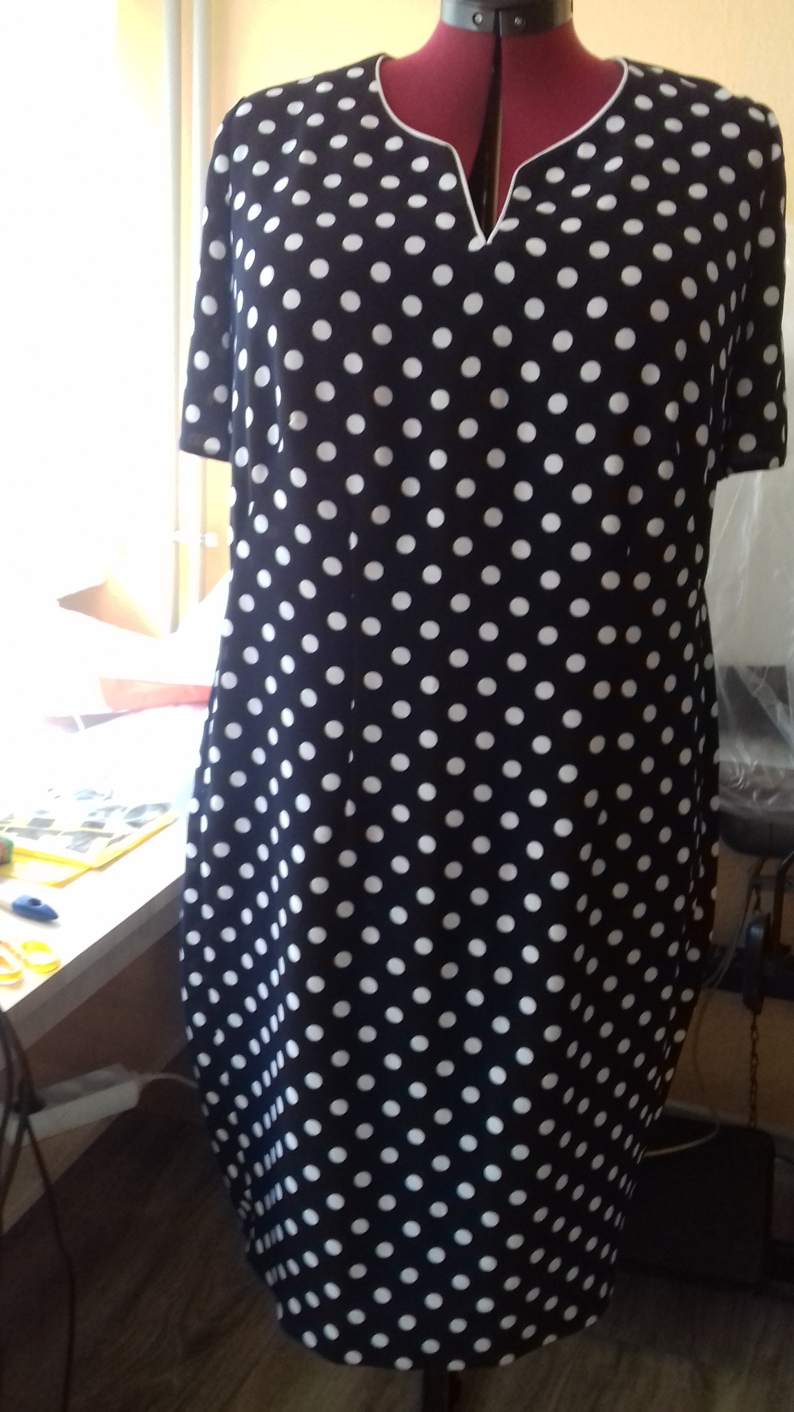 Sukienka w granatowo-białe grochy na czerwonym manekinie krawieckim, z widocznym stołem krawieckim w tle.