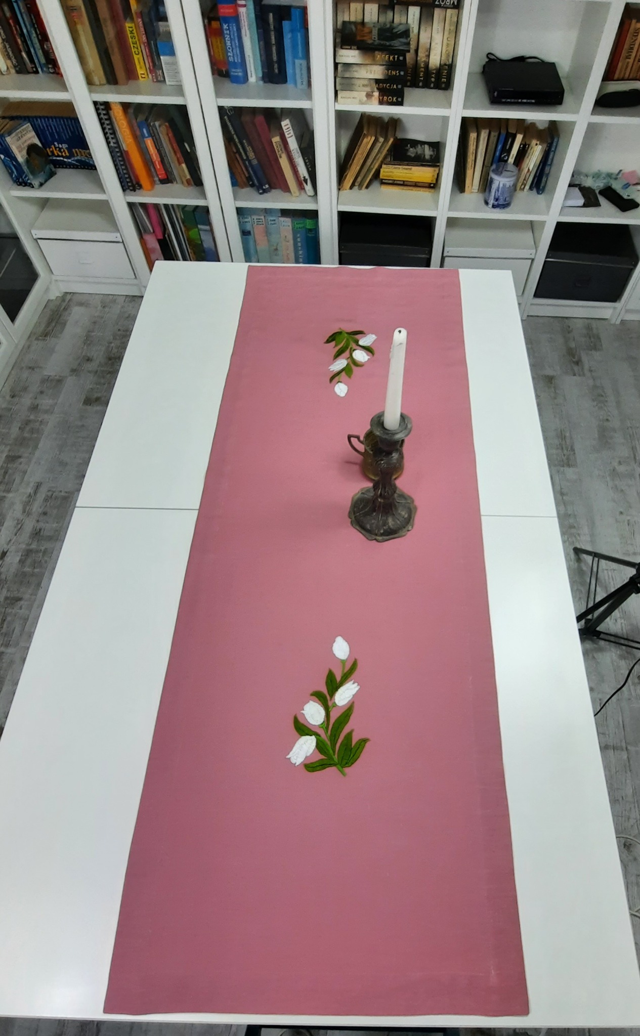 Różowy bieżnik z haftowanymi tulipanami i świecznikiem na białym stole, w tle regał z książkami.