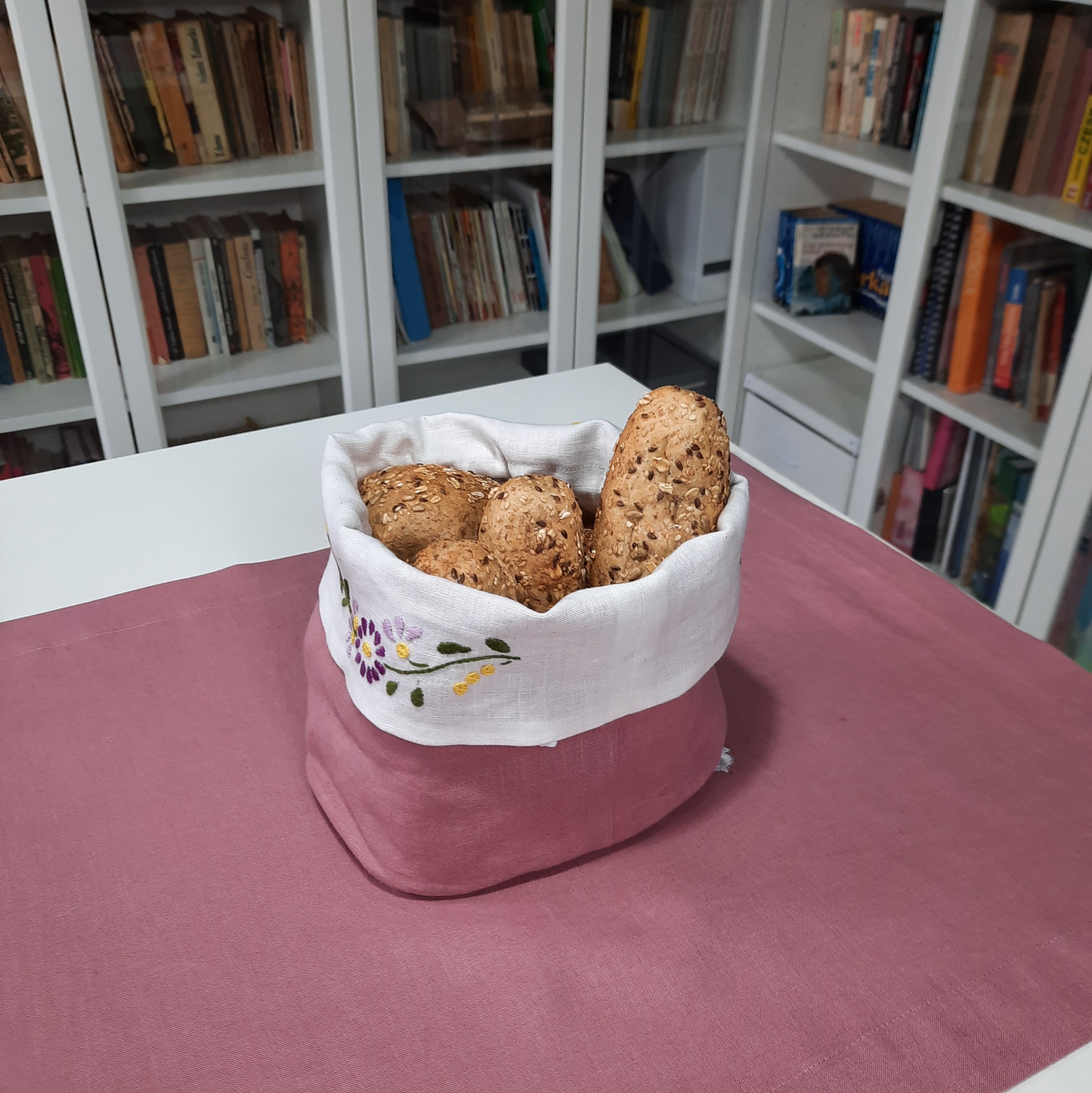 Ręcznie szyty, dwukolorowy koszyk na pieczywo z haftowanymi kwiatami, wypełniony bułkami z ziarnami, na tle białego regału z książkami.