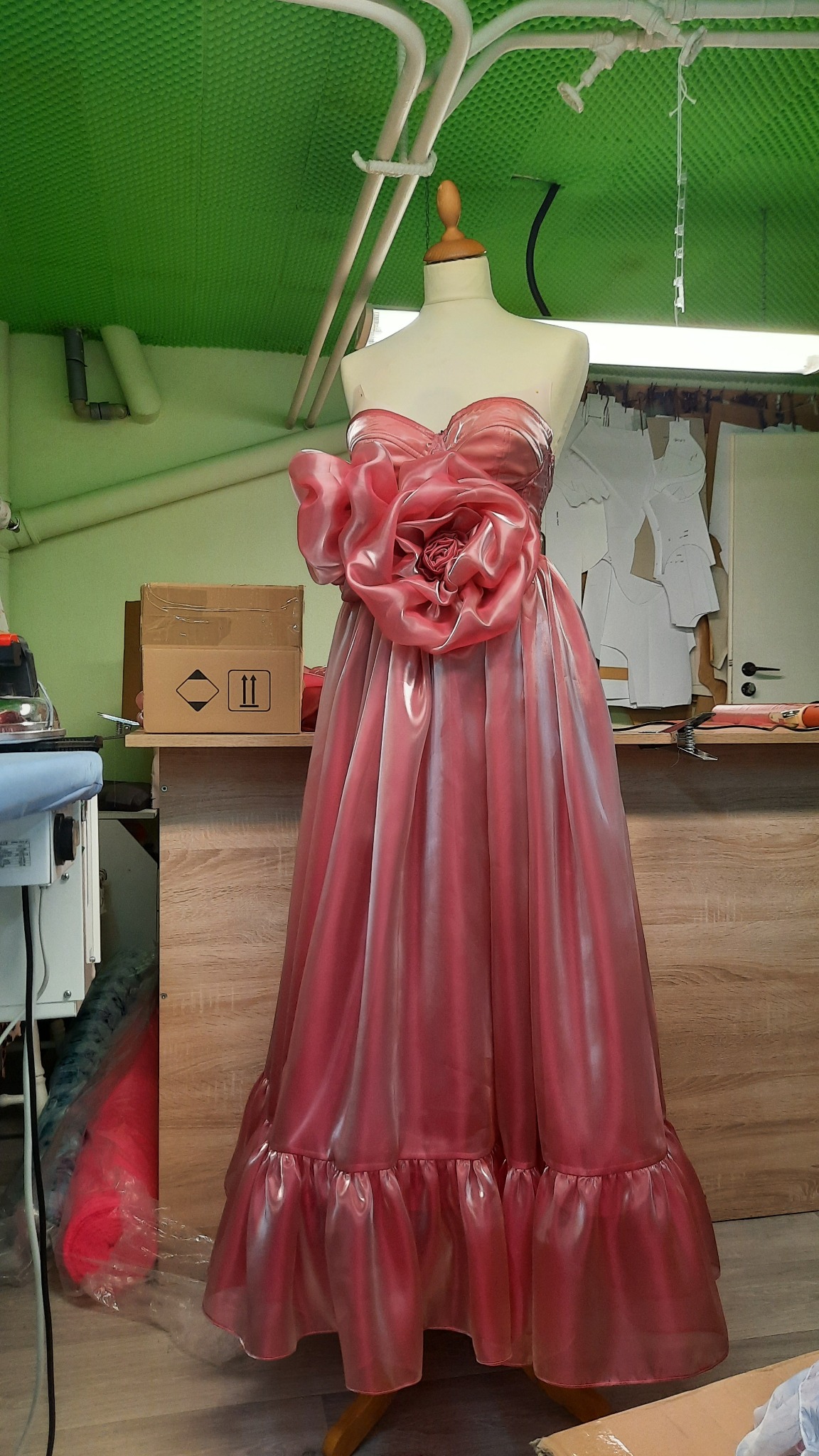 Różowa suknia balowa z dużą dekoracją kwiatową na manekinie, widoczne szablony odzieżowe w tle.