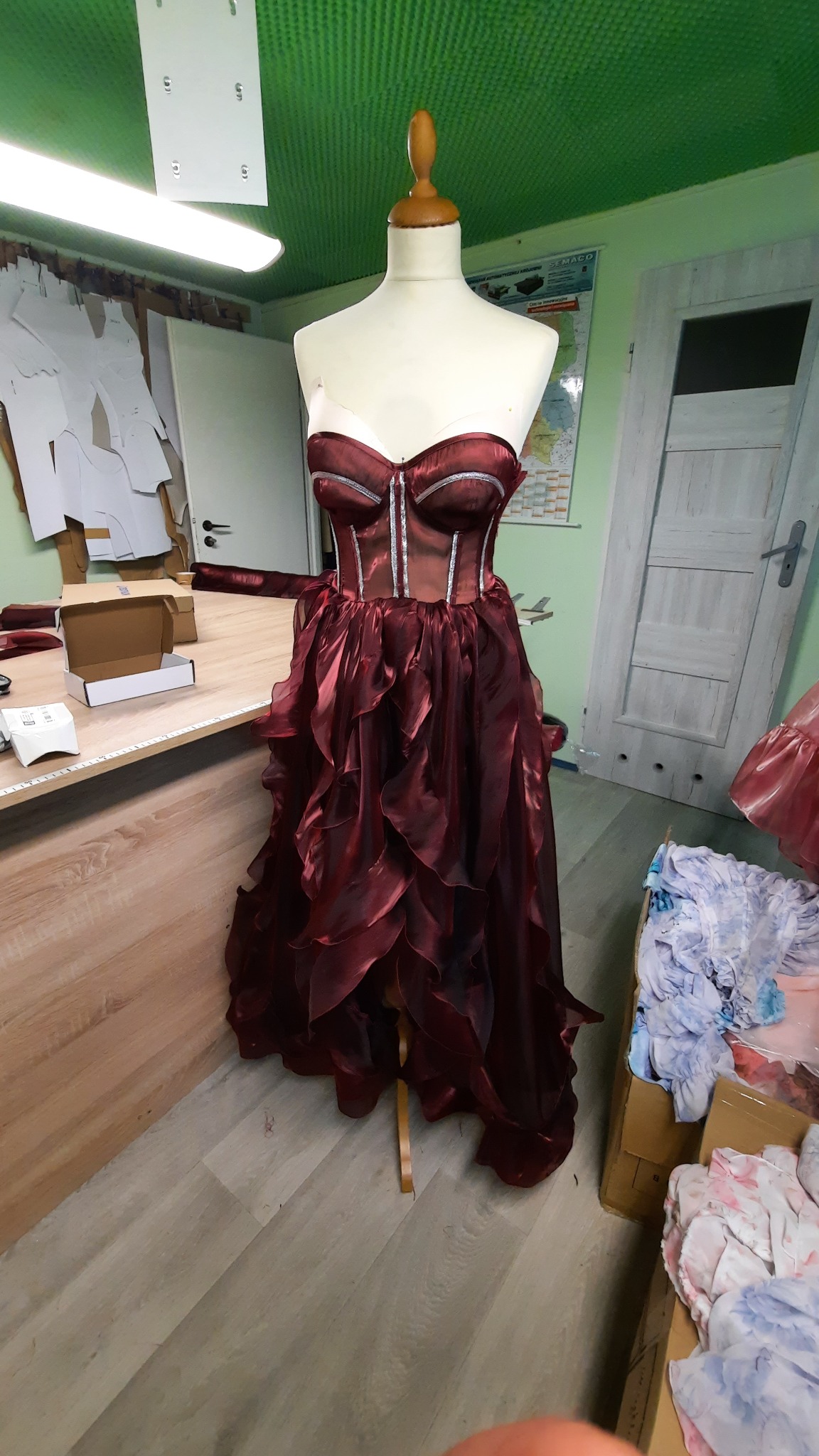 Burgundowa suknia wieczorowa z falbanami, prezentowana na manekinie w pracowni krawieckiej, obok wzory wykrojów i narzędzia.