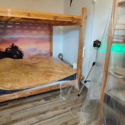 Wojtek Wojnarowski - Drewniana antresola z łóżkiem w trakcie montażu, zabezpieczona folią, poziomowana laserowo, widoczna tapeta z motywem tropikalnym i regał z książkami.