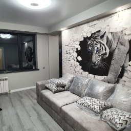 Dream_house_pl_ua - Szare wnętrze pokoju z tapetą 3D przedstawiającą białego tygrysa przebijającego się przez mur z cegieł, szara sofa z poduszkami zabezpieczonymi folią, czarne biurko i półki, szara podłoga imitująca...