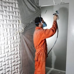 Dream_house_pl_ua - Pracownik w kombinezonie i masce maluje ścianę natryskowo na szaro, obok ściana z dekoracyjnymi panelami 3D, zabezpieczona folią malarską.