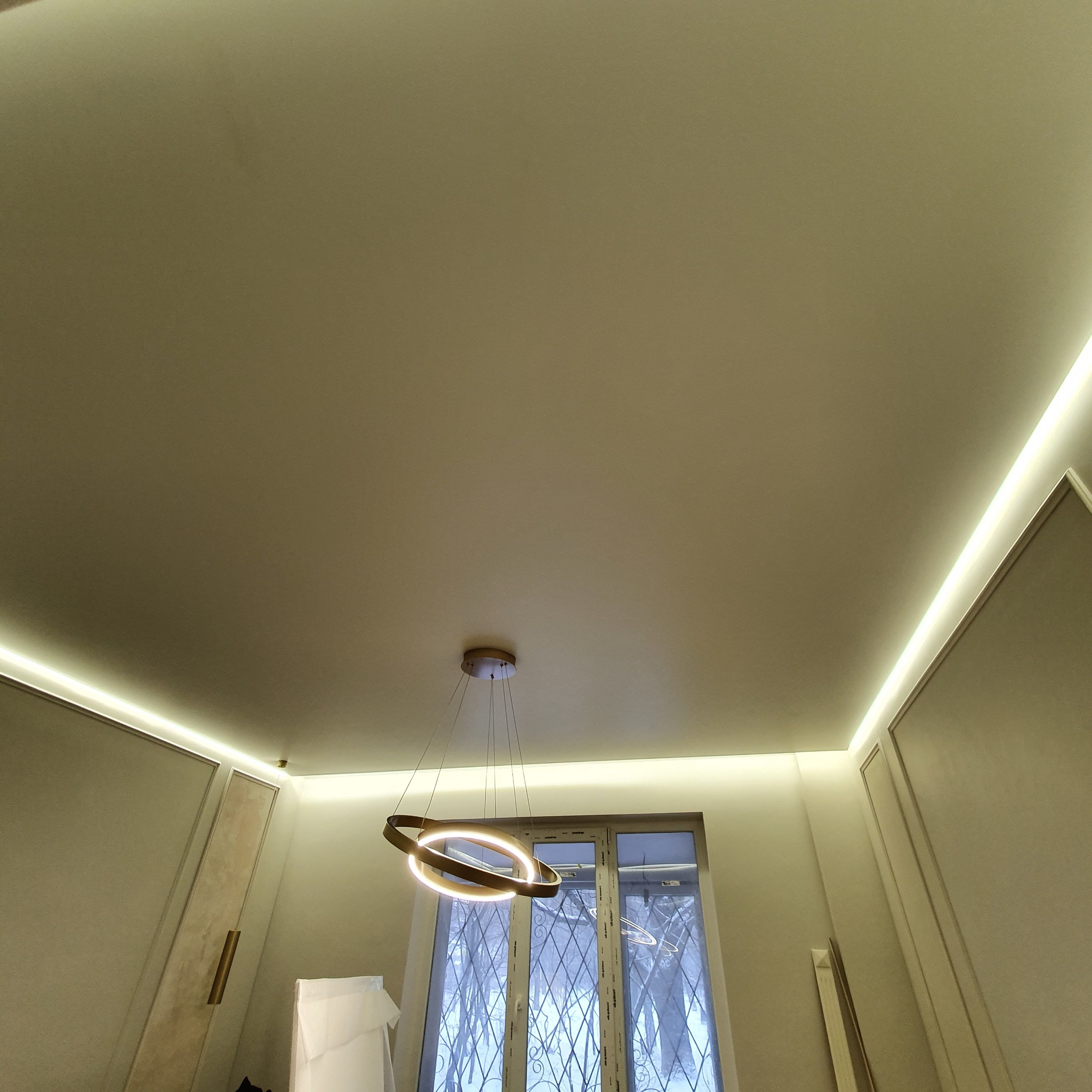 Nowoczesne oświetlenie sufitowe z listwami LED wzdłuż ścian i okrągłą lampą wiszącą na tle okna z kratami.