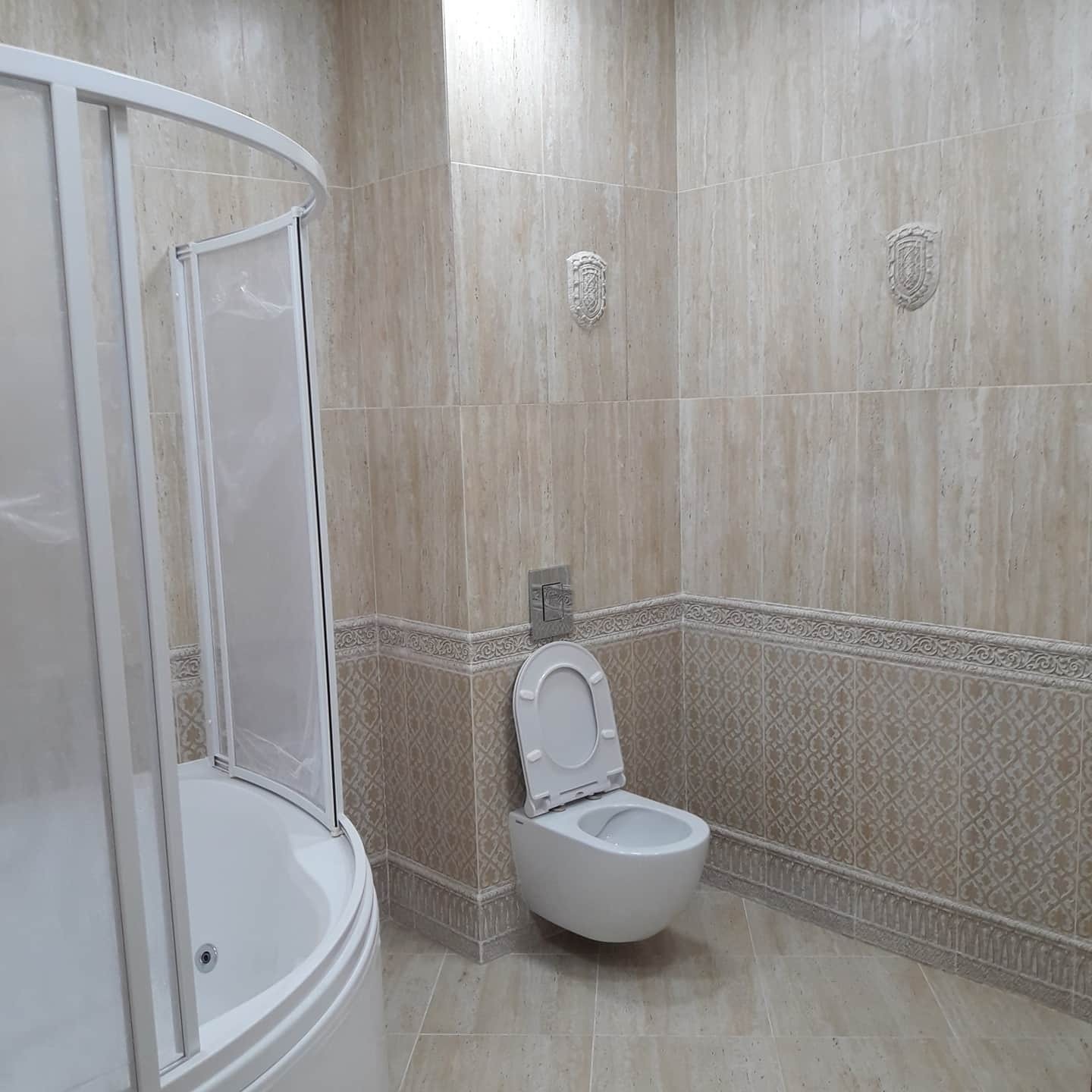 Wykończona łazienka w Poznaniu z beżowymi płytkami imitującymi trawertyn, dekoracyjnym pasem płytek z wzorem, wiszącą toaletą z otwartą klapą i kabiną prysznicową z zaokrąglonym frontem. Na ścianie...