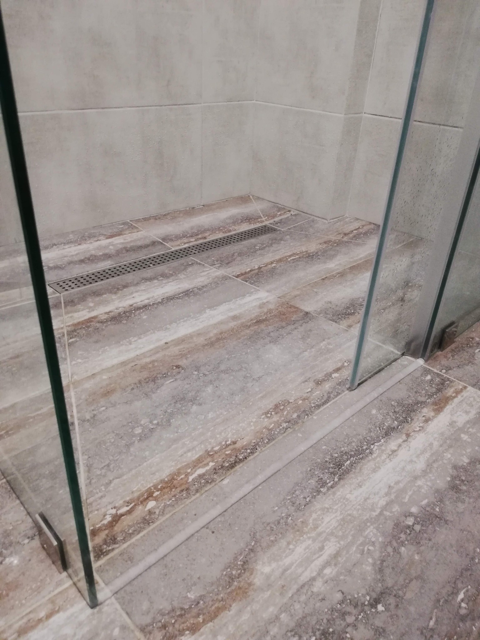 Kabina prysznicowa z odpływem liniowym w podłodze wyłożonej płytkami imitującymi drewno, widoczne mocowania szklanych ścianek.