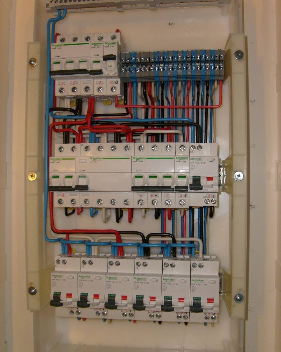 Rozdzielnica elektryczna z bezpiecznikami i przewodami w kolorach czerwonym, niebieskim i czarnym, zamontowana w plastikowej obudowie, widoczne oznaczenia producenta Schneider.