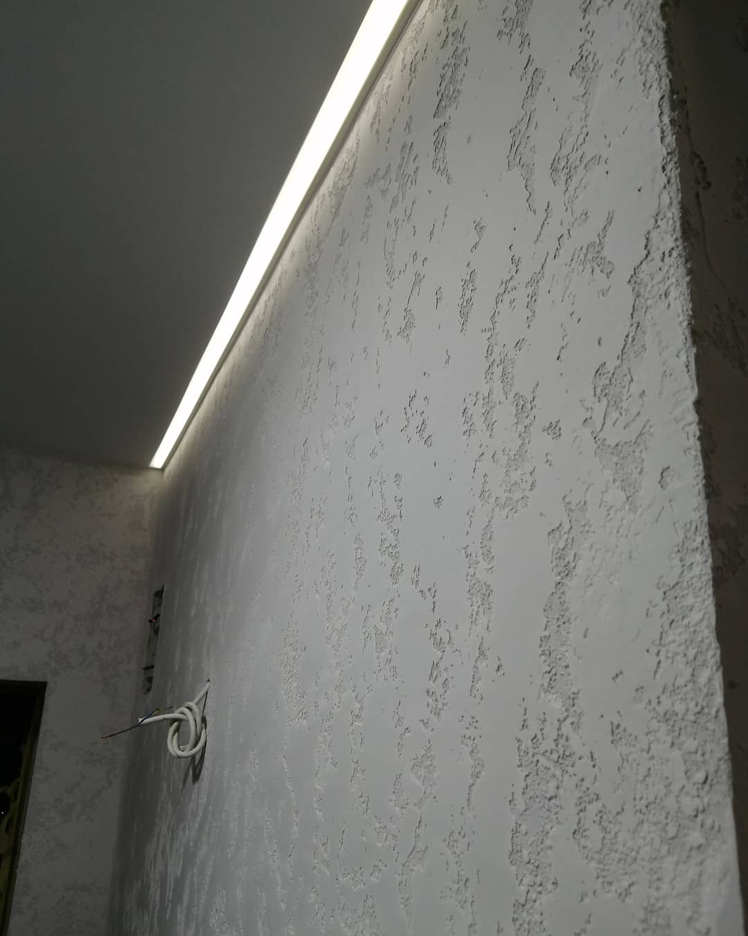 Ściana z dekoracyjnym tynkiem strukturalnym w kolorze szarym, oświetlona listwą LED pod sufitem; widoczne przewody elektryczne.