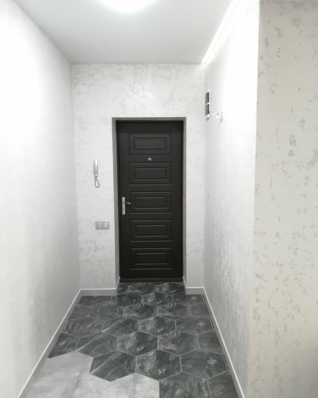 Wnętrze przedpokoju z ciemnymi drzwiami wejściowymi, szarą podłogą z heksagonalnych płytek imitujących marmur oraz ścianami o fakturze dekoracyjnego tynku w odcieniach bieli i szarości. Widoczny...