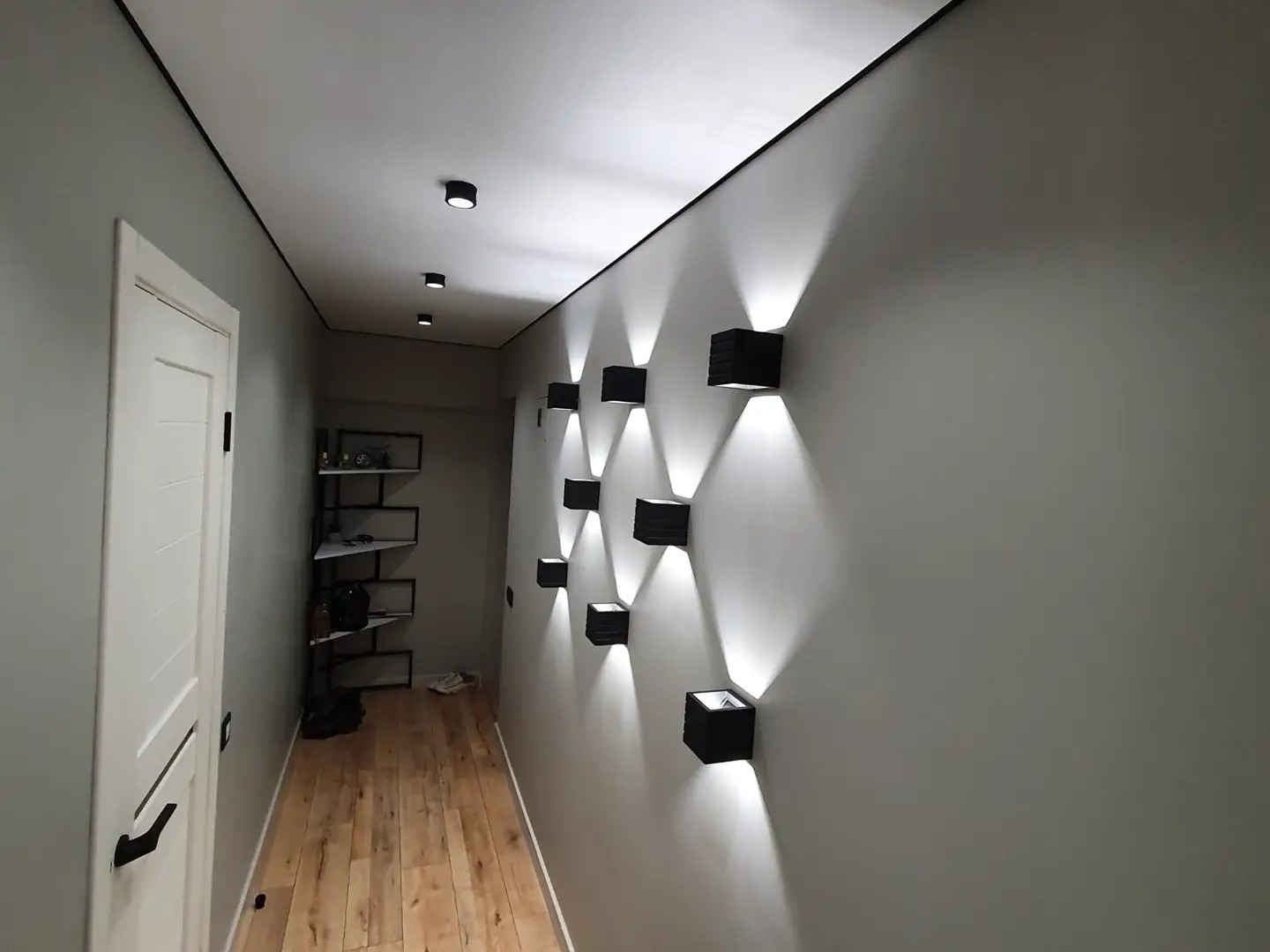 Korytarz z drewnianą podłogą i szarą ścianą, na której zamontowano rząd czarnych, kwadratowych lamp ściennych, emitujących jasne, rozproszone światło. Na końcu korytarza widoczny regał z półkami.