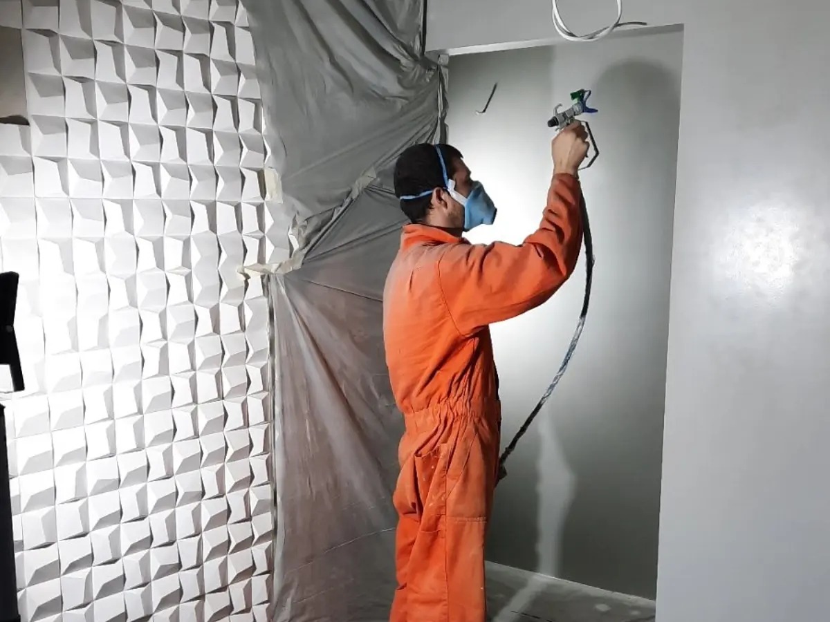 Pracownik w kombinezonie i masce maluje ścianę natryskowo na szaro, obok ściana z dekoracyjnymi panelami 3D, zabezpieczona folią malarską.
