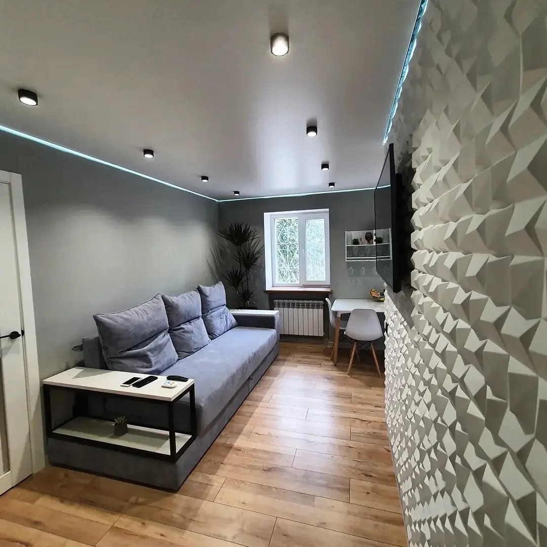 Nowoczesny salon z szarą sofą, stolikiem bocznym, trójwymiarową ścianą dekoracyjną, drewnianą podłogą i oknem z rośliną.