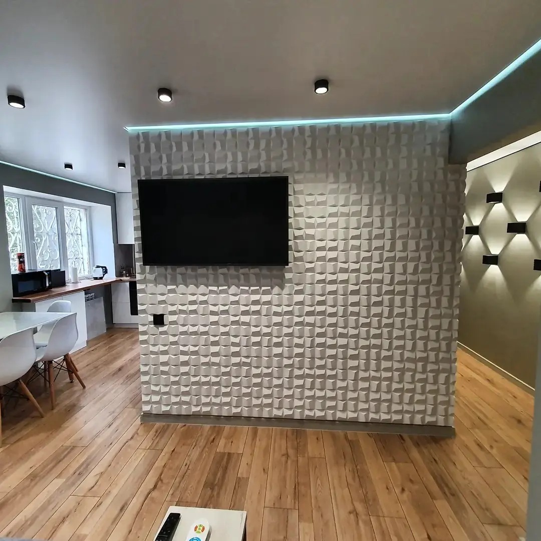 Nowoczesne wnętrze: ściana z trójwymiarowych paneli, podświetlenie LED, wiszący telewizor, parkiet, widoczny fragment kuchni z białymi meblami.