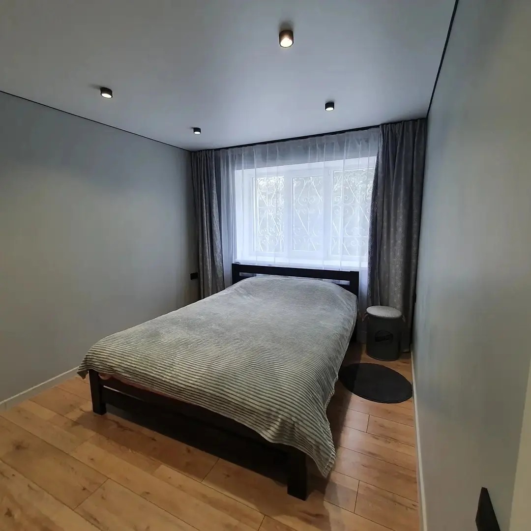 Nowoczesna sypialnia z drewnianą podłogą, szarą pościelą w paski i szarymi ścianami, oświetlona punktowymi lampami sufitowymi i światłem dziennym przez okno z białymi firankami i szarymi zasłonami.