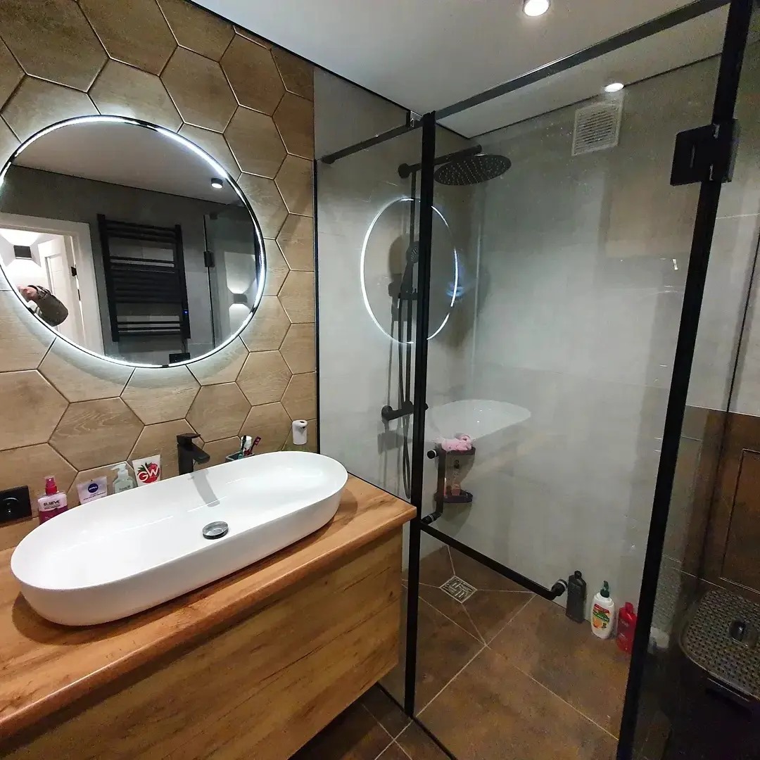 Nowoczesna łazienka z heksagonalnymi płytkami imitującymi drewno, owalną umywalką nablatową na drewnianym blacie i kabiną prysznicową typu walk-in z czarnymi profilami.