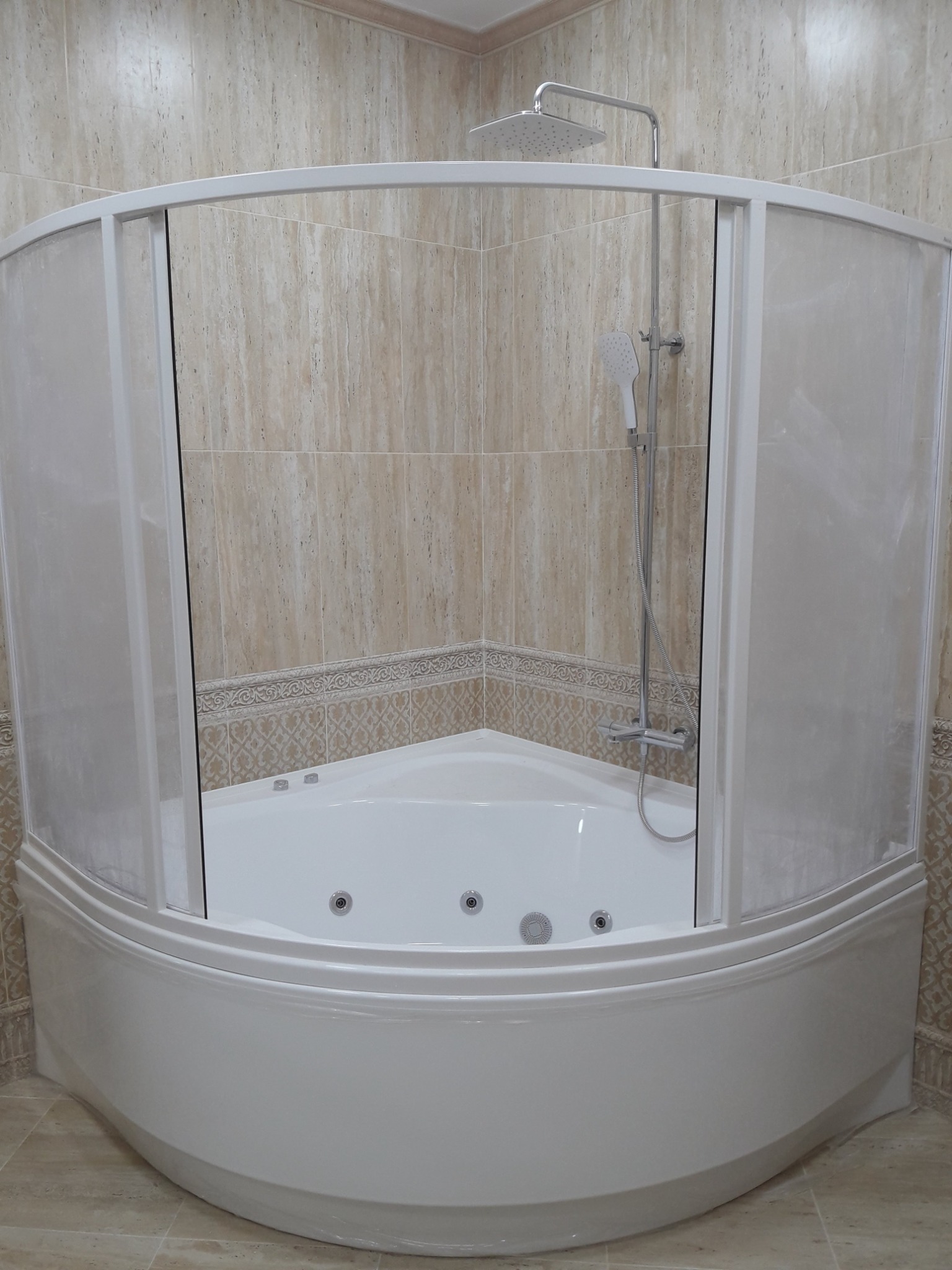 Biała, narożna wanna z hydromasażem i przeszkloną kabiną prysznicową, zamontowana w łazience wyłożonej beżowymi płytkami z delikatnym wzorem.