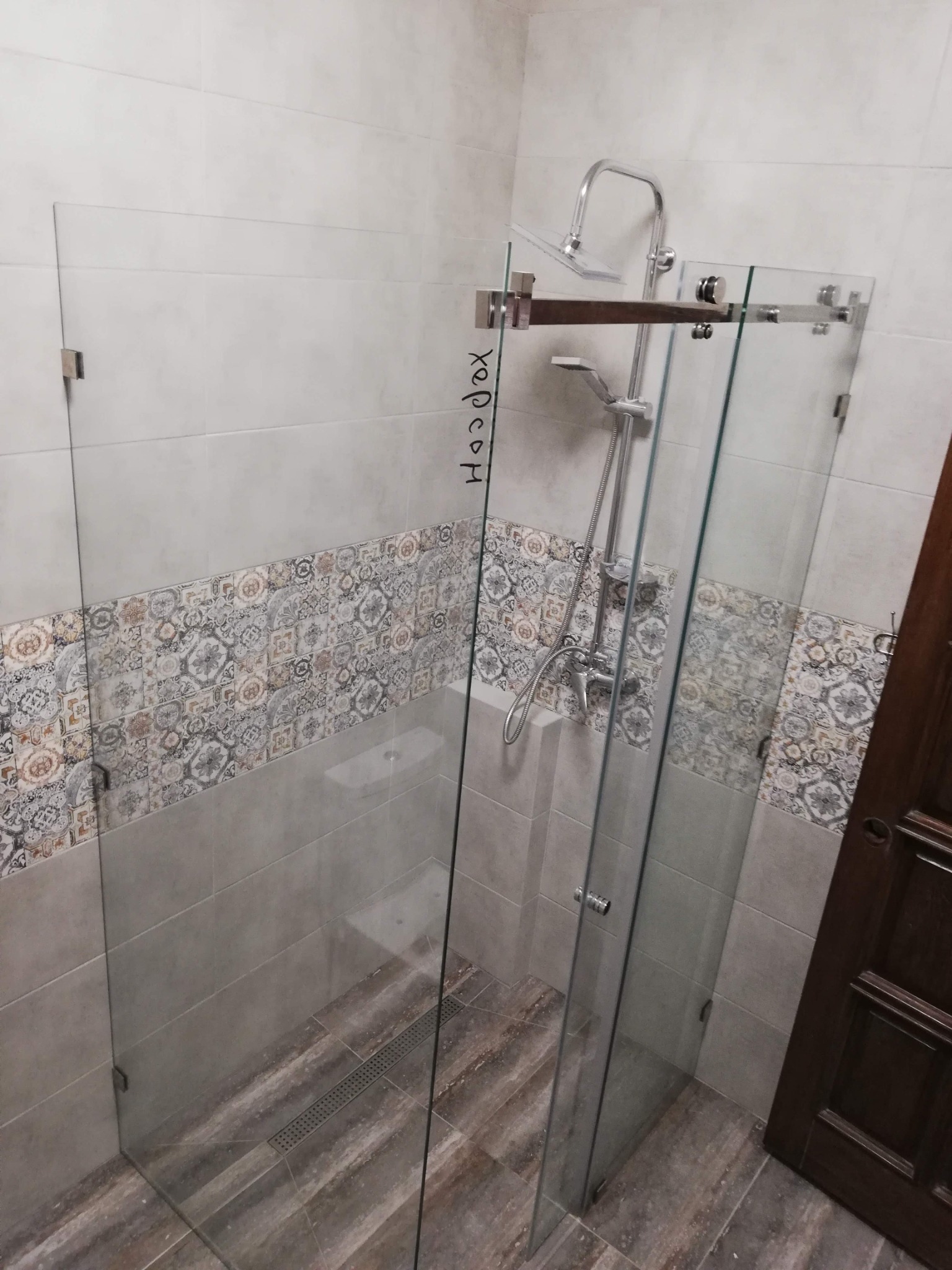 Nowoczesna kabina prysznicowa ze szklanymi ściankami, chromowaną armaturą i odpływem liniowym, widoczna częściowo napisana na szkle nazwa 'Hardox'.