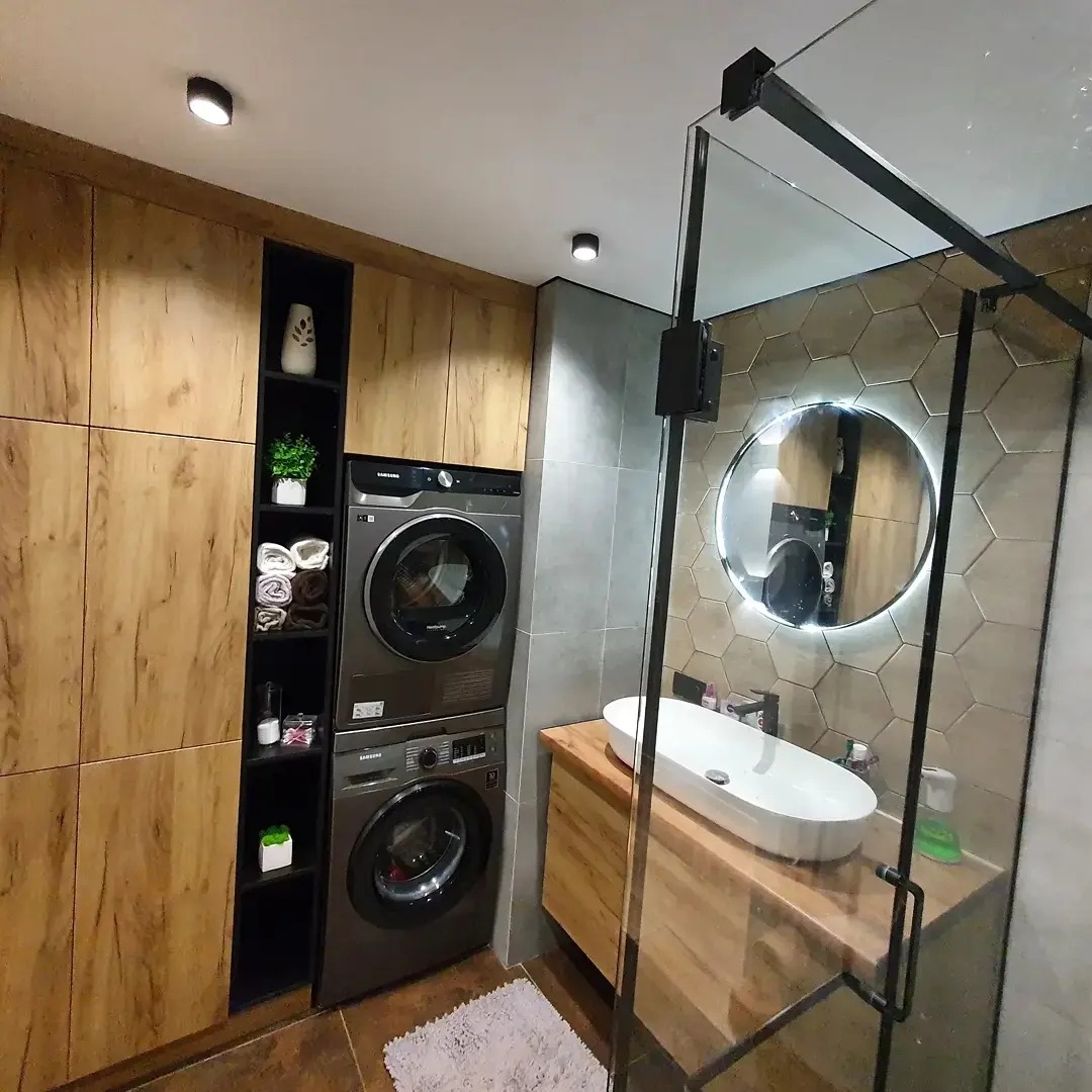 Nowoczesna łazienka z zabudowaną pralką i suszarką w drewnianej szafce, umywalka nablatowa na drewnianej szafce z podświetlanym lustrem w kształcie koła oraz kabina prysznicowa z czarnymi profilami.