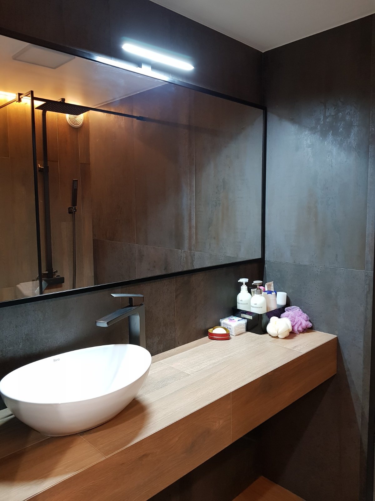 Nowoczesna łazienka z umywalką nablatową Rea, baterią w kolorze grafitu i dużym lustrem z oświetleniem LED. Na blacie drewnopodobnym stoją kosmetyki i akcesoria.