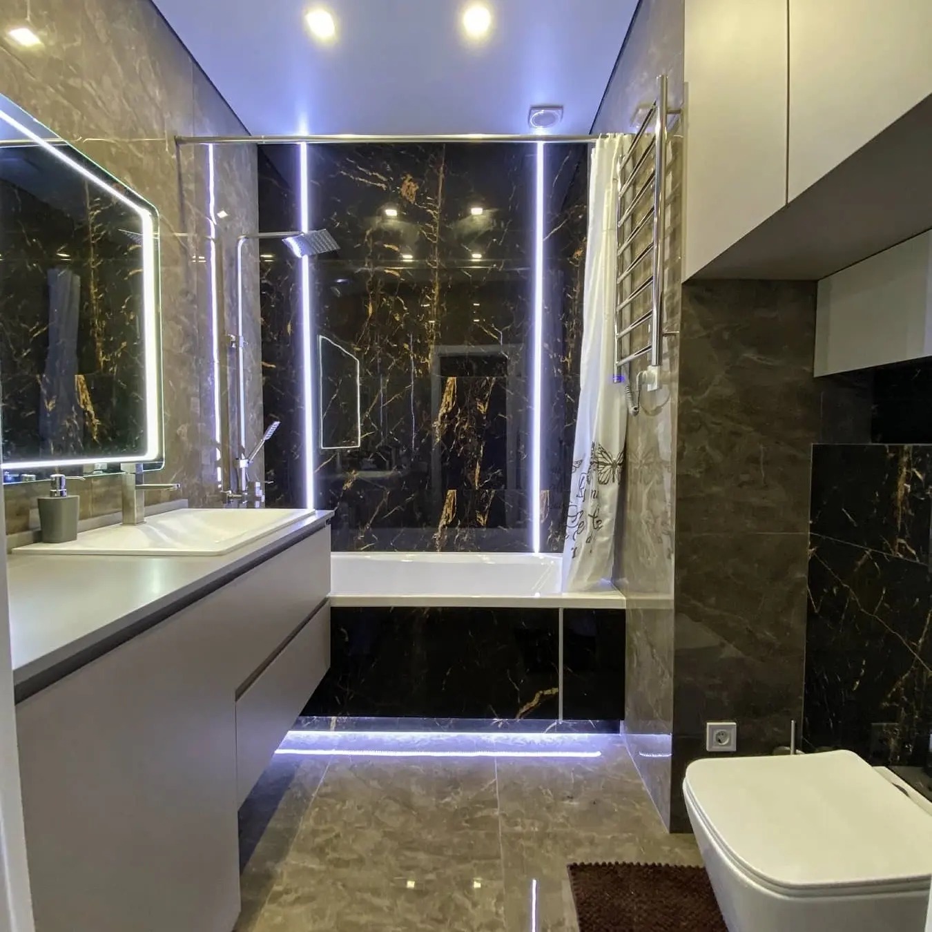 Nowoczesna łazienka z czarnymi marmurowymi płytkami, podświetleniem LED za wanną i pod szafką umywalkową, oraz grzejnikiem drabinkowym na ścianie.